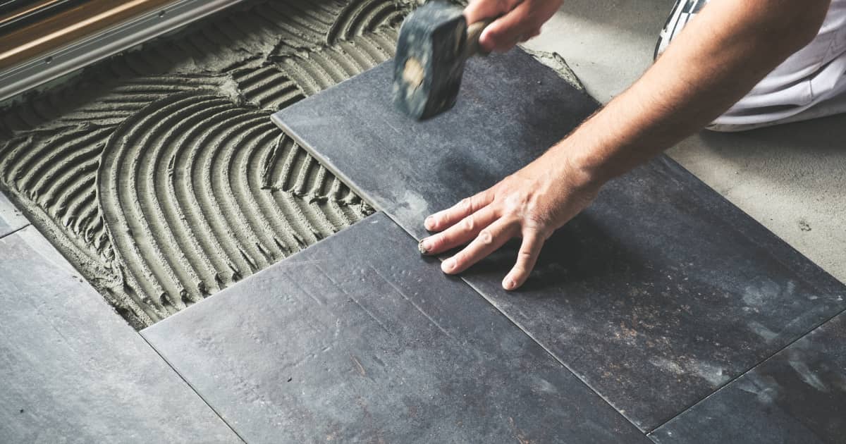 Pose de carrelage au sol dans une cuisine, un artisan ajuste une dalle en grès cérame gris foncé.