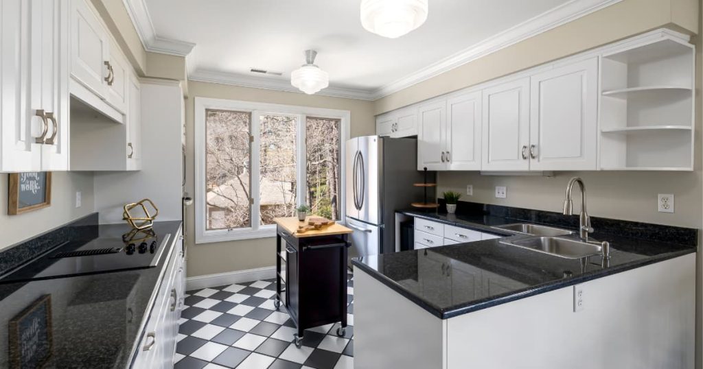 Cuisine lumineuse avec meubles blancs, plan de travail en granit noir et sol en carrelage noir et blanc à damier.
