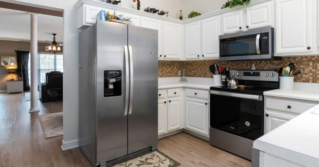 Cuisine moderne avec armoires blanches, réfrigérateur en inox, four et micro-ondes intégrés, plan de travail clair et crédence en carreaux mosaïque marron.