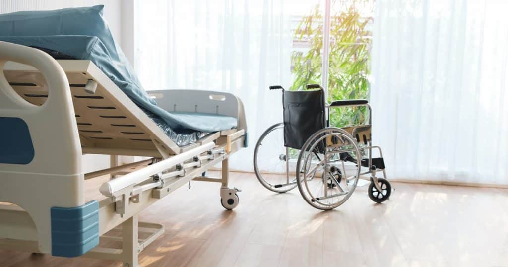 Un lit simple au dossier redressé à côté d'un fauteuil roulant devant une baie vitrée ensoleillée dans une chambre d'hôpital. 