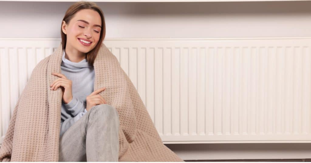 Une femme qui sourit adossée sur un chauffage avec une couverture sur le dos