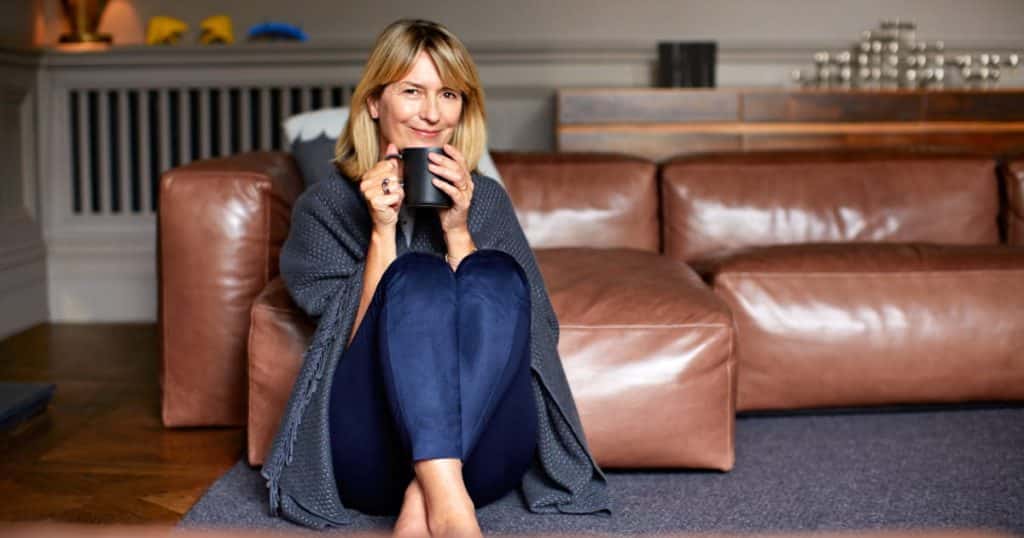 Une femme assise sur son tapis qui sourit, une couverture sur le dos et un mug en mains 