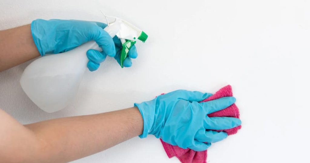 Mains gantées qui nettoient un mur blanc un vaporisateur dans une main et un torchon dans l’autre.