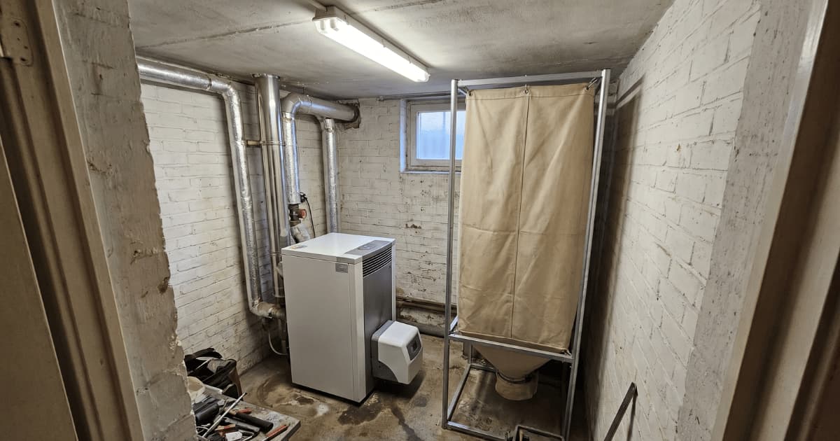 Une chaudière à pellets compacte blanche est installée dans une petite cave aux murs de briques blanchis à côté d'un silo textile beige sur cadre métallique avec des conduites d'évacuation en aluminium au plafond et un éclairage fluorescent.
