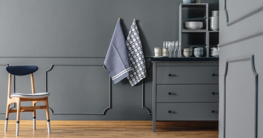 Cuisine avec un mur gris et un buffet assorti, des verres et de la vaisselles sont rangés dans le meuble, des torchons sont accrochés au mur et une chaise à côté.