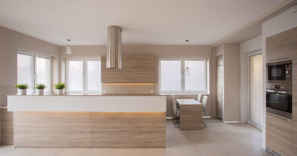 Cuisine moderne avec meubles en bois clair, îlot central lumineux, hotte en inox, coin repas intégré et grandes fenêtres laissant entrer la lumière naturelle.