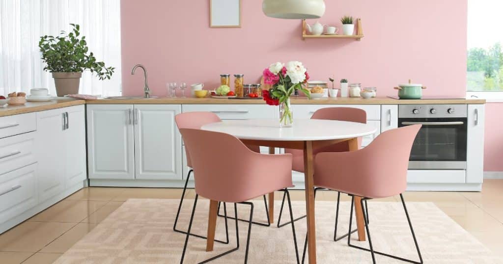 Une cuisine blanche moderne avec un four et un mur rose pastel, une table ronde au milieu entourée de chaines avec un vase avec des fleurs.