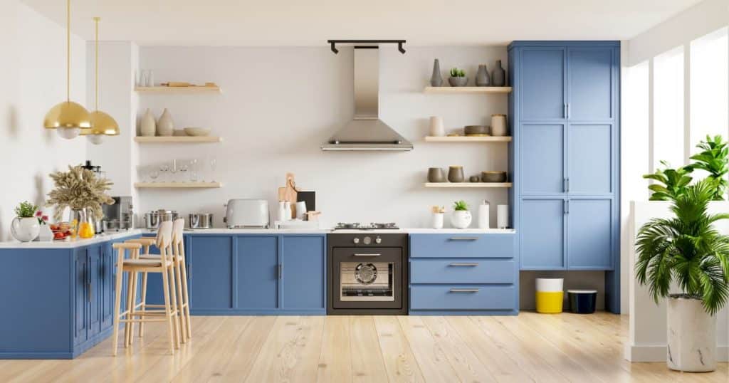Une cuisine blanche aux meubles bleus, deux chaises, deux suspensions dorées, un four, une hôtte, des ustensiles de cuisine, un toaster, des étagères en bois et des plantes