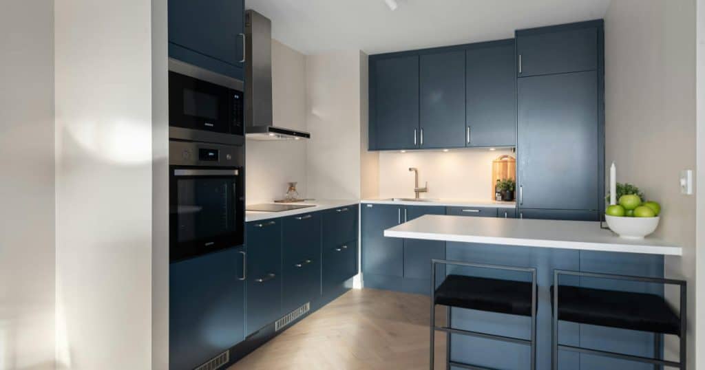 Cuisine avec des armoires bleu foncé mates, un plan de travail blanc, des électroménagers encastrés en inox, une hotte murale, et un coin bar avec deux tabourets noirs. Un bol de pommes vertes et une bougie décorent le plan de travail.