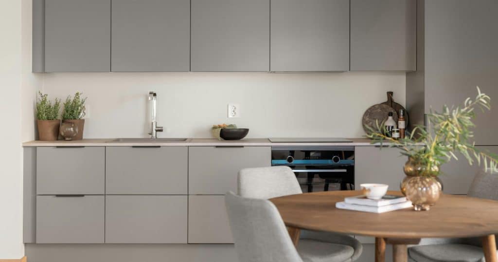 Cuisine minimaliste gris clair avec façades sans poignées, plan de travail en béton et table en bois avec décoration végétale.
