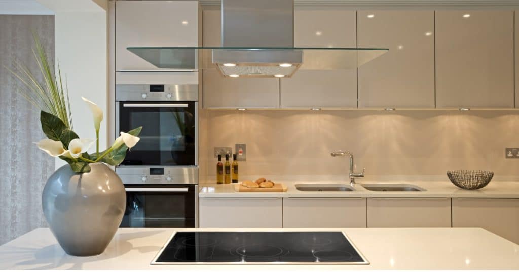 Cuisine moderne beige avec îlot central, plaque de cuisson vitrocéramique, hotte en verre, double évier inox, four encastré et grand vase décoratif avec fleurs blanches.