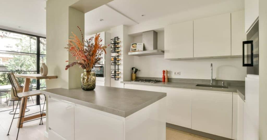 Cuisine moderne blanche avec îlot central, plan de travail en gris, hotte en inox, rangement mural à bouteilles et grande baie vitrée donnant sur l’extérieur.
