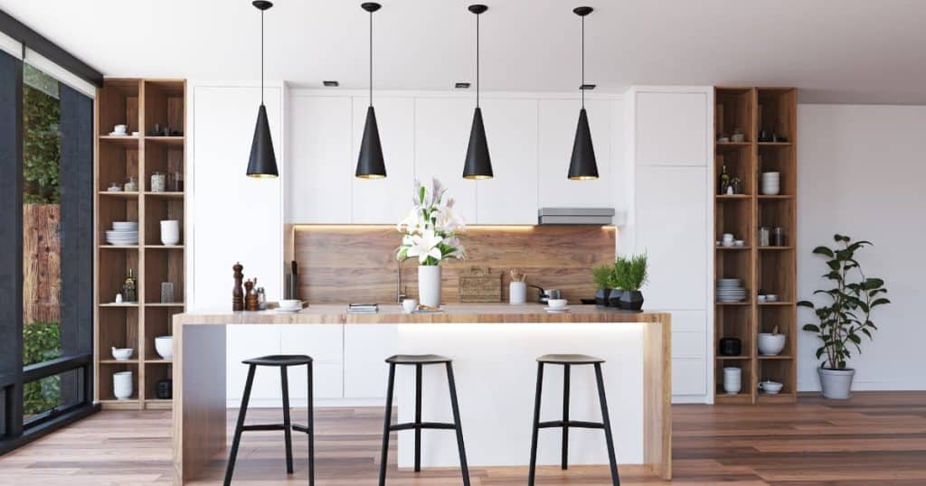 Une cuisine blanche avec des suspensions noires au dessus de l'ilot central avec trois chaises et des pots de plantes