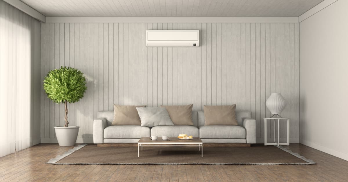 Salon avec un fauteuil à l’arrière plan, à droite un grand arbuste, à gauche un meuble avec une lampe. En hauteur il y a un climatiseur blanc.
En moyen plan il y a un table très basse.