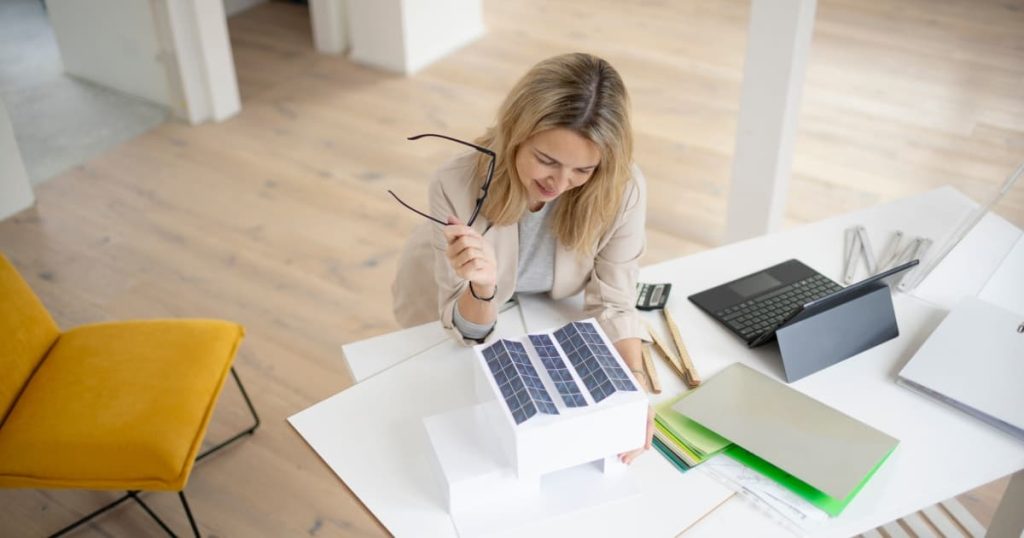 femme blonde qui tient des lunettes et regarde un bureau avec ordinateur, maquette de maison avec panneaux solaires