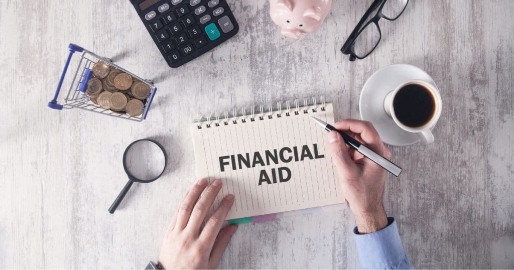 Table avec un carnet ligné où il est écrit "financial aid". Autour, il y a une loupe, un caddie avec des sous, une calculatrice, un cohonnet, une paire de lunette de vue et un café.