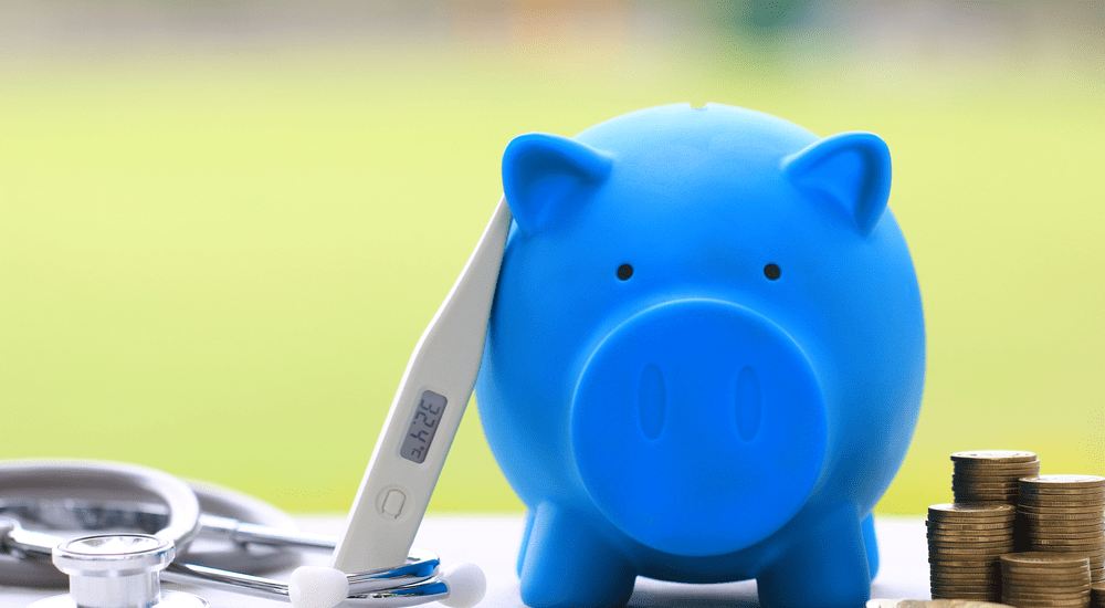 Un thermomètre médical à côté d'un stéthoscope repose sur une tirelire bleue en forme de cochon, à côté de pièces de monnaie empilées. 