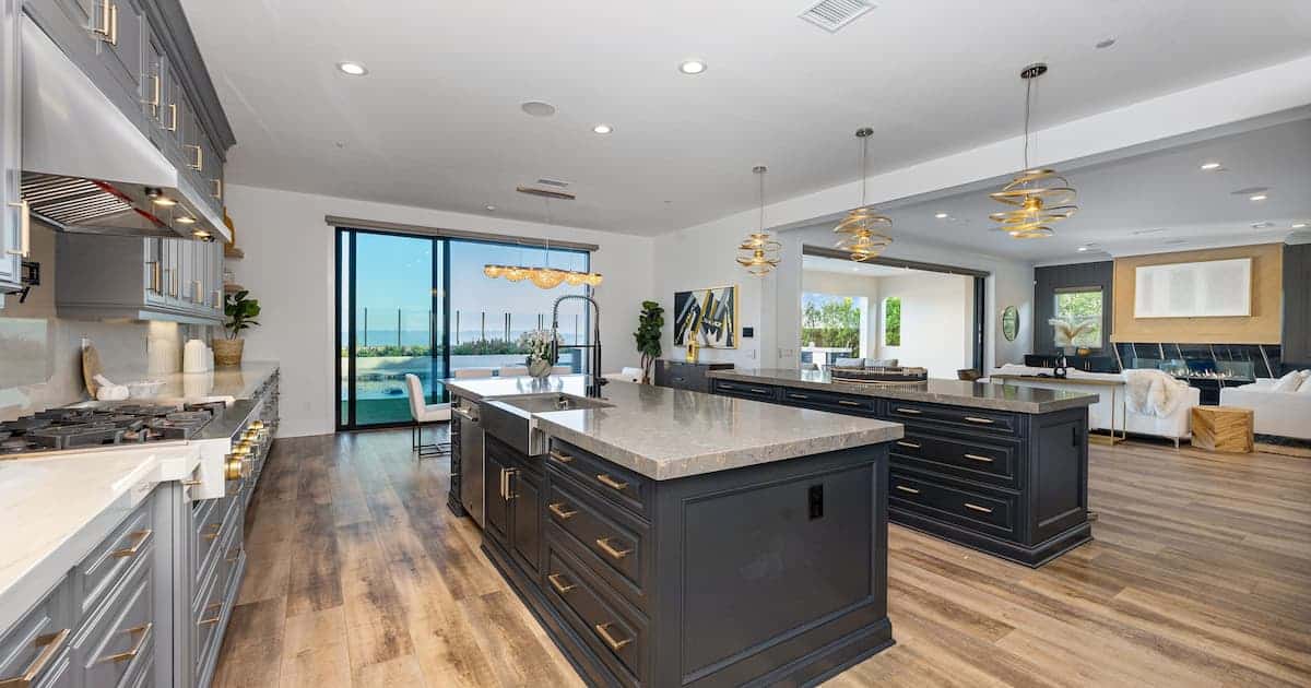 Cuisine moderne à aire ouverte avec armoire sombre, îlot et éclairage suspendu, menant à un salon spacieux avec de grandes portes vitrées.