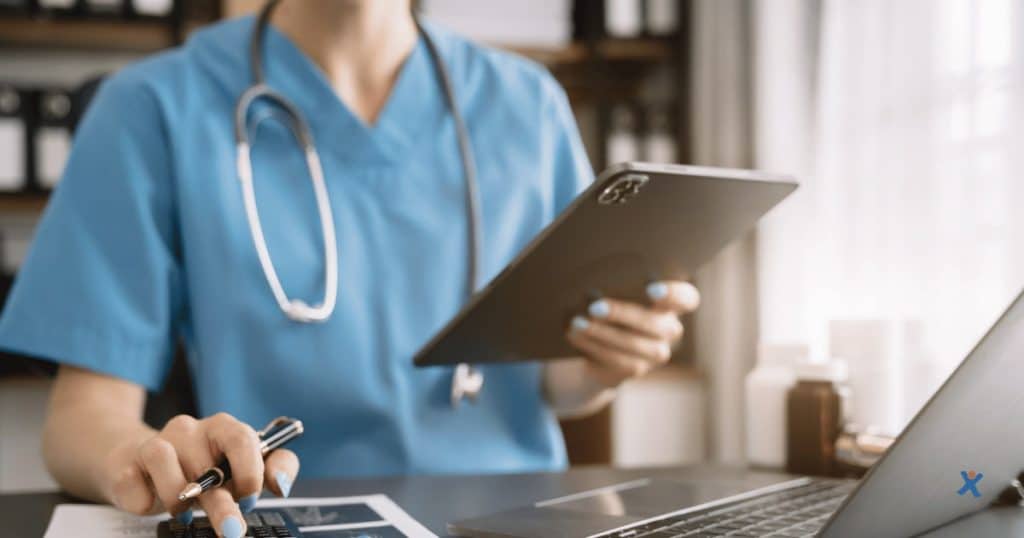 Infirmière en uniforme bleu consultant des données sur une tablette tout en prenant des notes au bureau, avec un stéthoscope autour du cou.