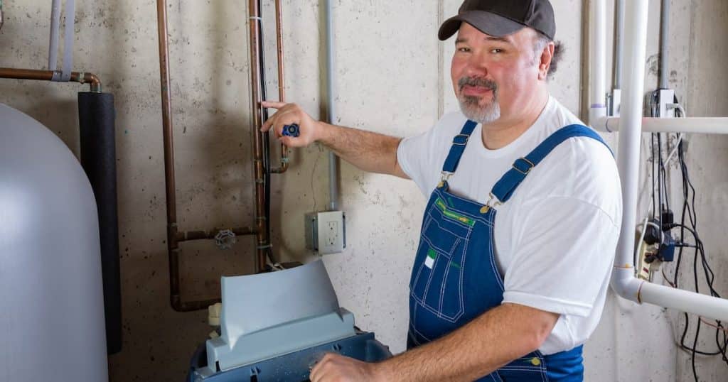 Ouvrier souriant en salopette installant ou travaillant sur un adoucisseur d'eau dans une buanderie, se tournant pour sourire à l'appareil photo.