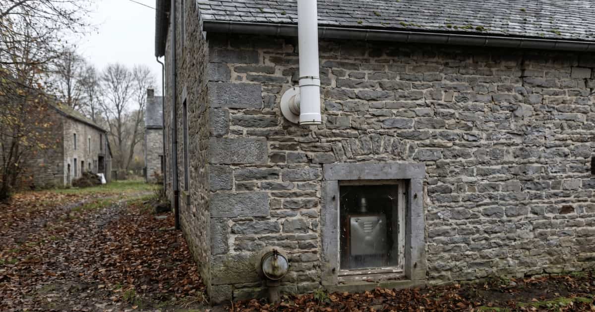 Un conduit d'évacuation blanc en PVC longe la façade en pierre grise d'une maison wallonne tandis qu'un bouchon de remplissage de mazout rouillé est visible près du sol sous une petite fenêtre de cave par temps d'automne couvert.