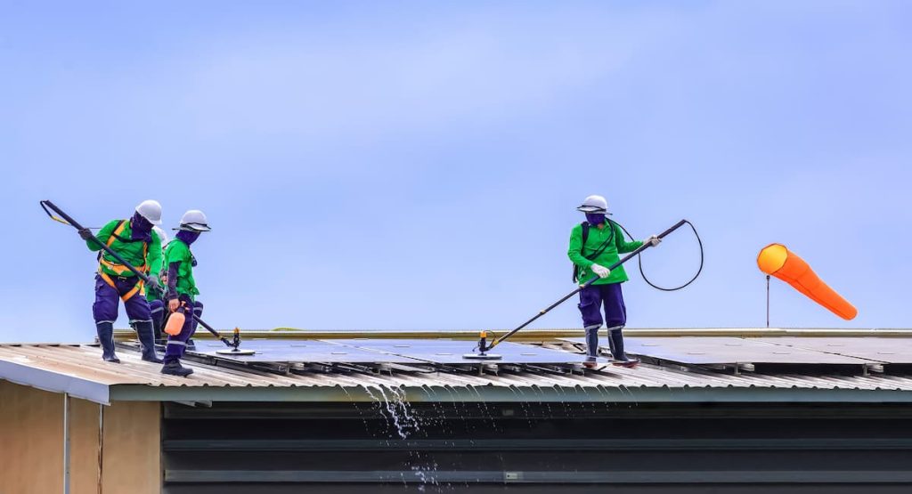 Nettoyage de panneau solaire par des professionnels : 3 hommes avec perche télescopique en train de nettoyer les panneaux solaires sur le toit d'un bâtiment