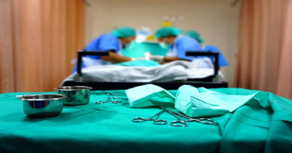 table d'une salle d'opération avec des ciseaux de chirurgien et du matériel médical et en arrière plan un patient en train de se faire opérer