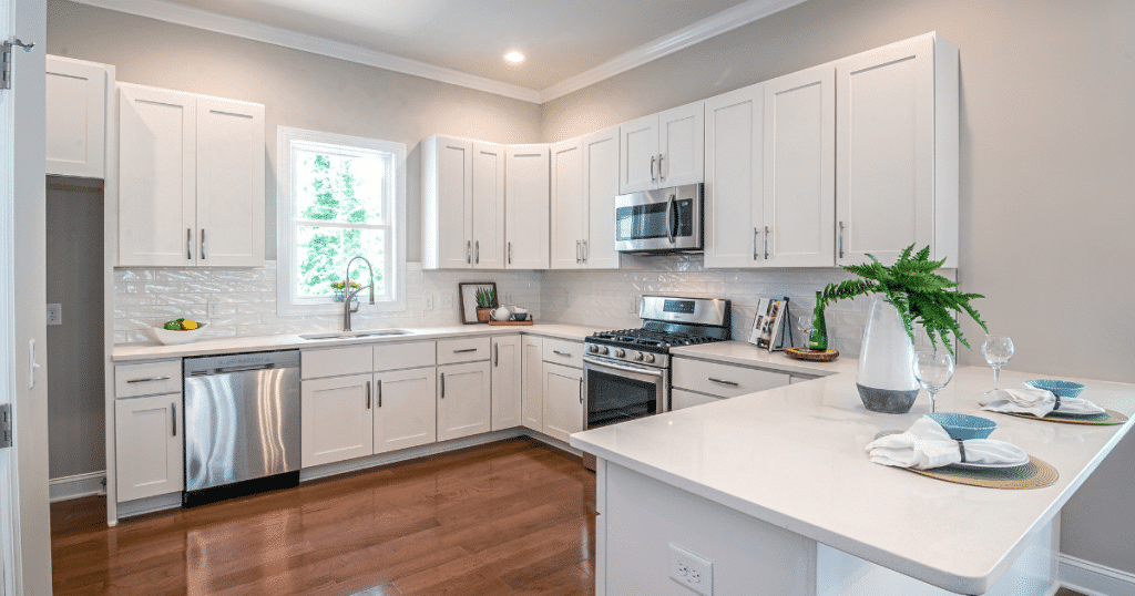 Cuisine moderne lumineuse avec armoires blanches, plan de travail en quartz, électroménagers en inox et sol en bois, incluant un îlot avec décoration de vaisselle et plante verte.