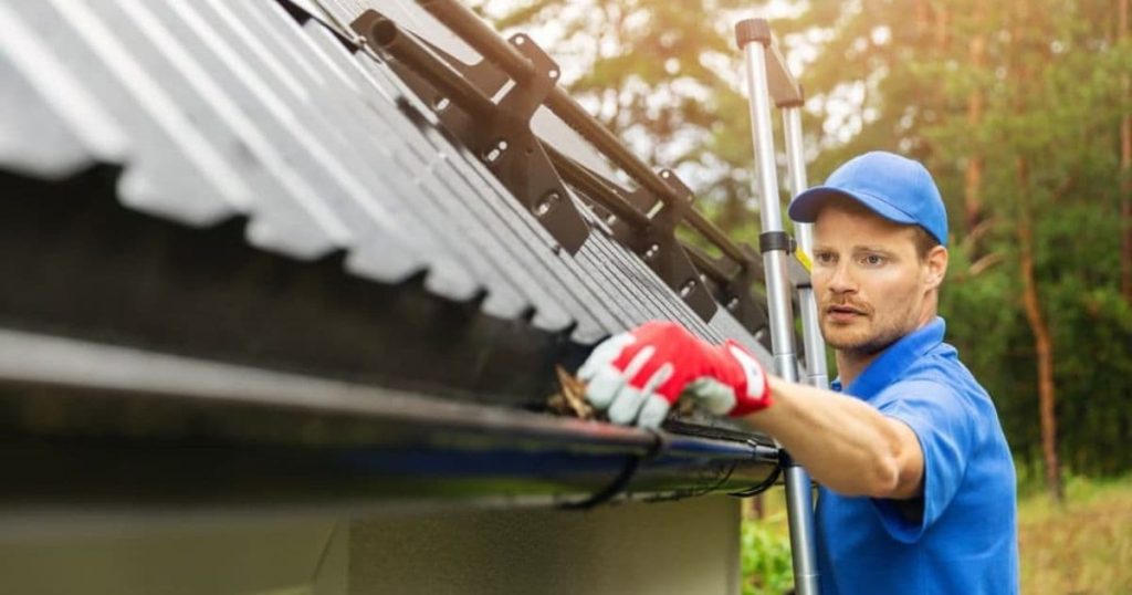 réparateur sur une échelle qui répare une gouttière avec une casquette bleue et des gants rouges 