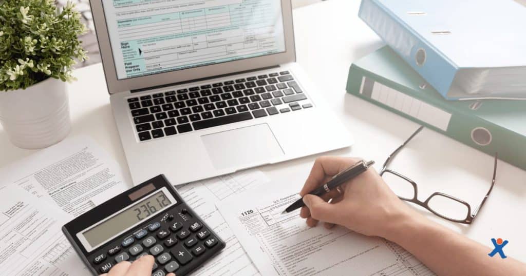 Un comptable ou un entrepreneur examine des documents fiscaux et encode des données à l’aide d’un ordinateur portable et d’une calculatrice. La scène montre un bureau organisé avec des classeurs, des formulaires d’impôts, des lunettes et une plante décorative. L’image illustre une situation de travail comptable ou fiscal.