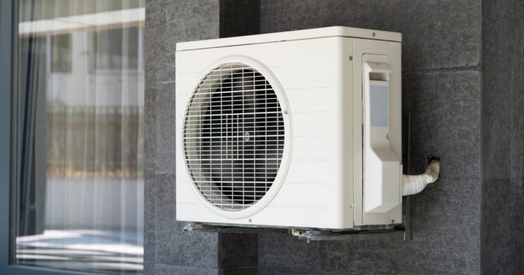 Une pompe à chaleur à l'extérieur d'un batiment gris a coté d'une fenêtre.