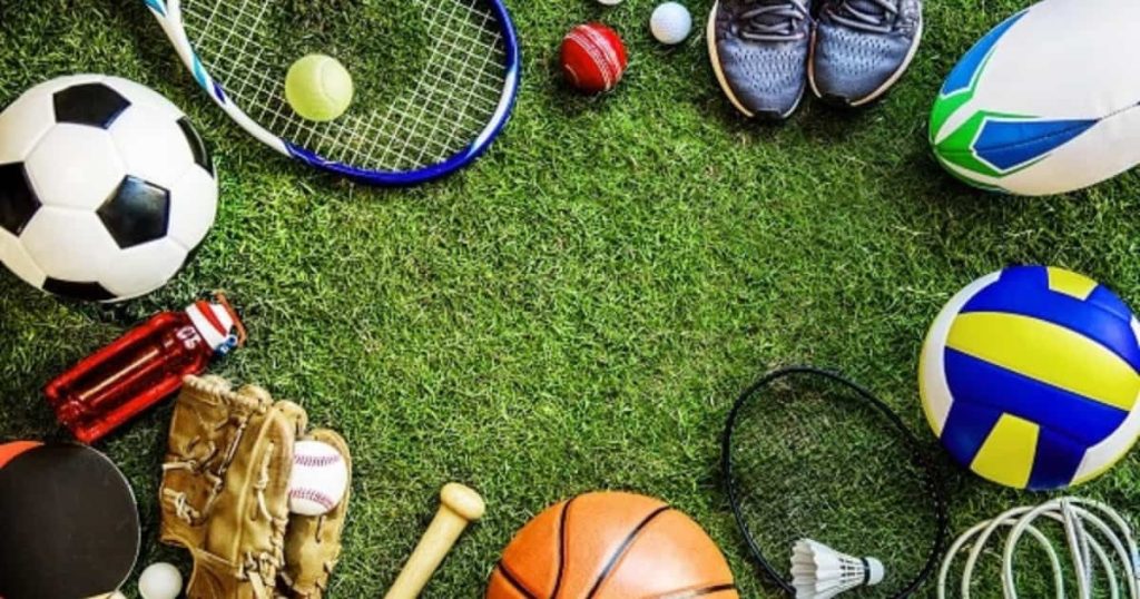 Vue de dessus de divers équipements sportifs disposés sur de l'herbe, incluant ballon de football, raquette de tennis, ballon de basket, gants de baseball, ballon de volleyball, chaussures de sport et gourde