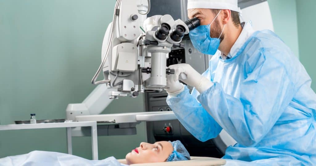 Un ophtalmologiste en tenue chirurgicale effectue une opération oculaire à l’aide d’un microscope spécialisé. Une patiente est allongée sur une table d’examen, regardant vers le haut, prête pour l’intervention.