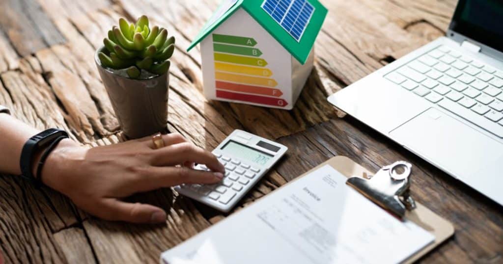 Une main utilise une calculatrice à côté d'une maison miniature avec étiquette énergétique