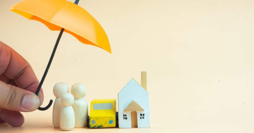 Main tenant un parapluie jaune au-dessus de figurines représentant une famille, une voiture et une maison en bois, symbolisant la protection offerte par une assurance