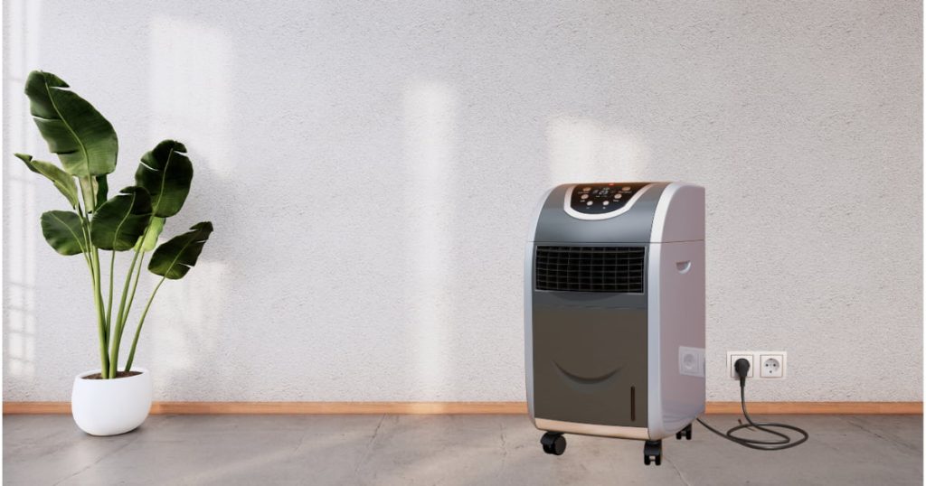 Climatiseur mobile gris foncé branché à une prise murale, posé sur un sol en béton dans une pièce moderne avec une plante verte en pot