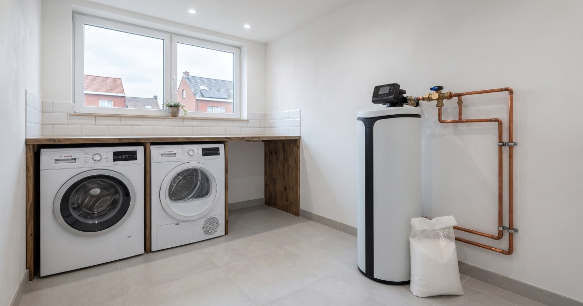 Un adoucisseur d'eau au sel blanc avec écran de contrôle digital est raccordé à des tuyaux en cuivre dans une buanderie moderne aux murs blancs, avec un sac de pastilles de sel posé au sol à côté de l'appareil et un lave-linge et sèche-linge encastrés sous un plan de travail en bois à gauche.