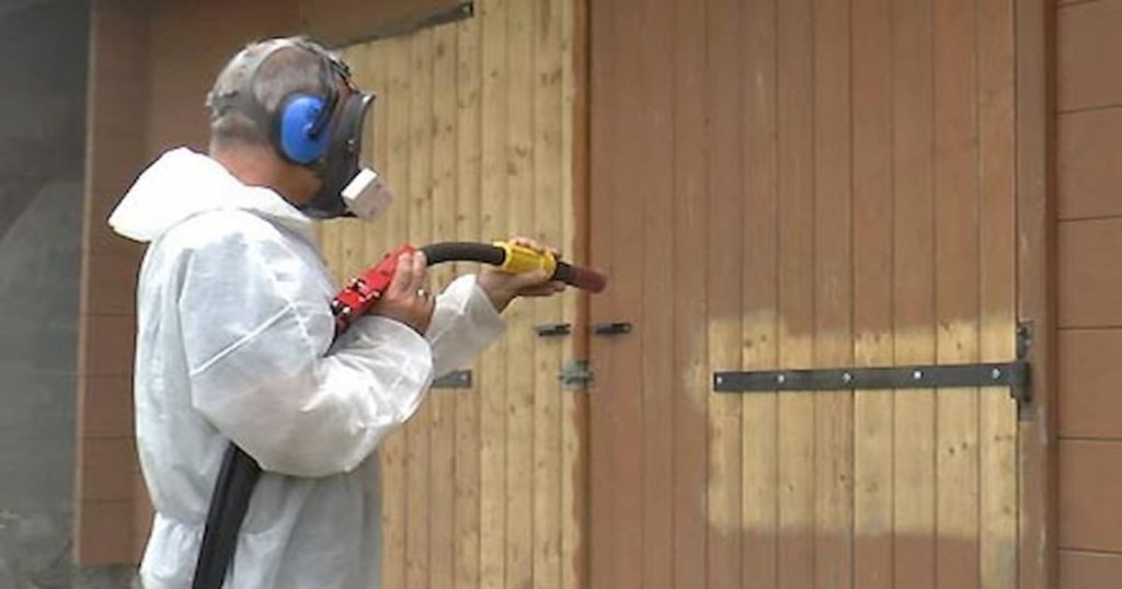Homme en tenue blanche utilisant une aérogommeuse sur une porte en bois