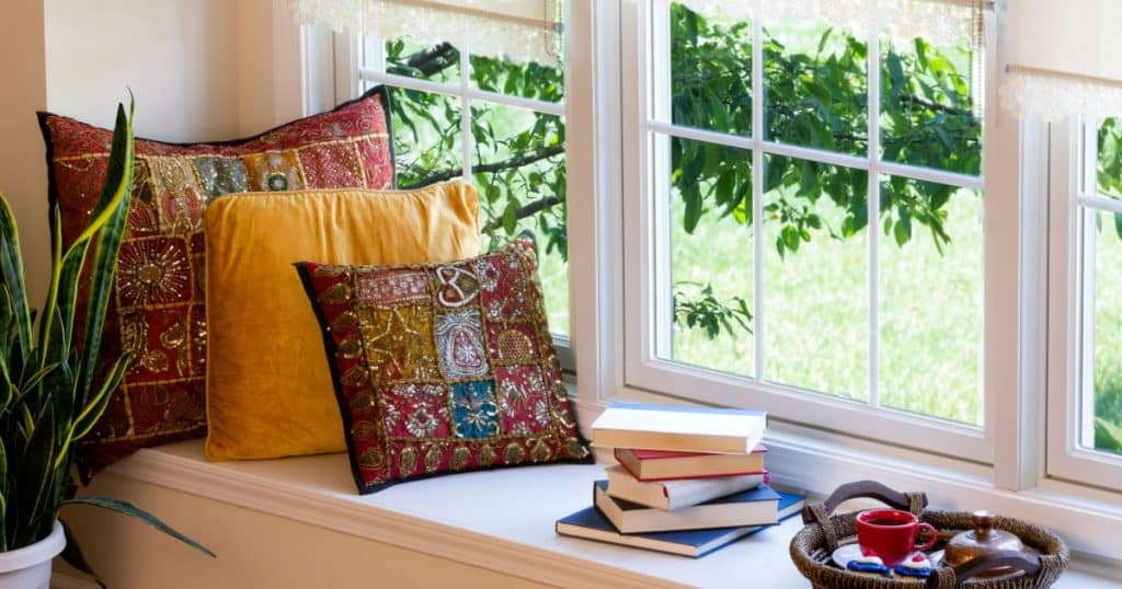 Vue d'un coin lecture dans une petite véranda, avec des coussins, des livres et une plante.