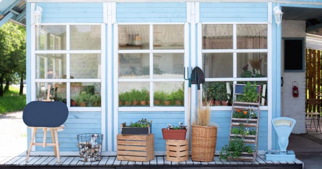Vue depuis l'extérieur d'une petite véranda attenante à une maison, on voit à travers les vitres des plants d'herbes aromatiques en pot. 