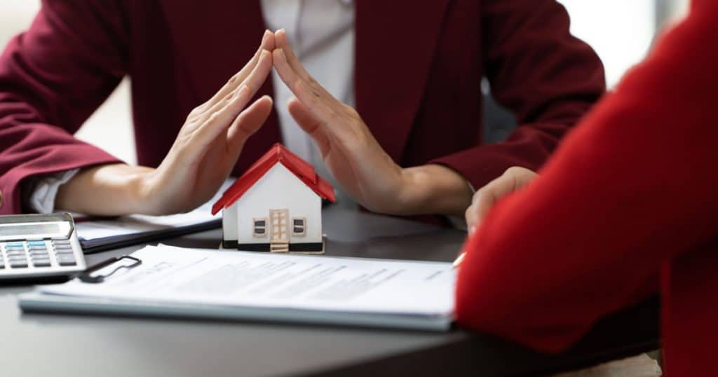 Une conseillère en assurance face à une cliente tient ses deux mains en toit au-dessus d'une maison miniature posée sur une table, à côté d'une calculatrice et de papiers. 