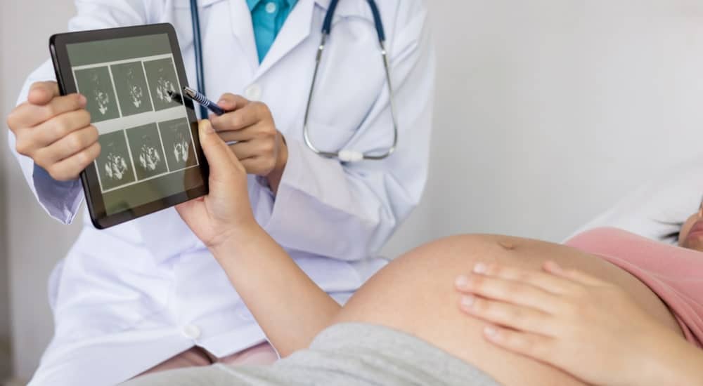 Femme enceinte couchée sur un lit d'hôpital qui découvre (sur une tablette) les échographies réalisées par un médecin.
