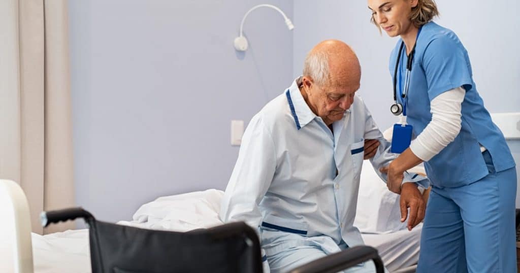 Une aide-soignante aide un homme âgé à se lever de son lit d'hôpital pour s'asseoir sur un fauteuil roulant. 