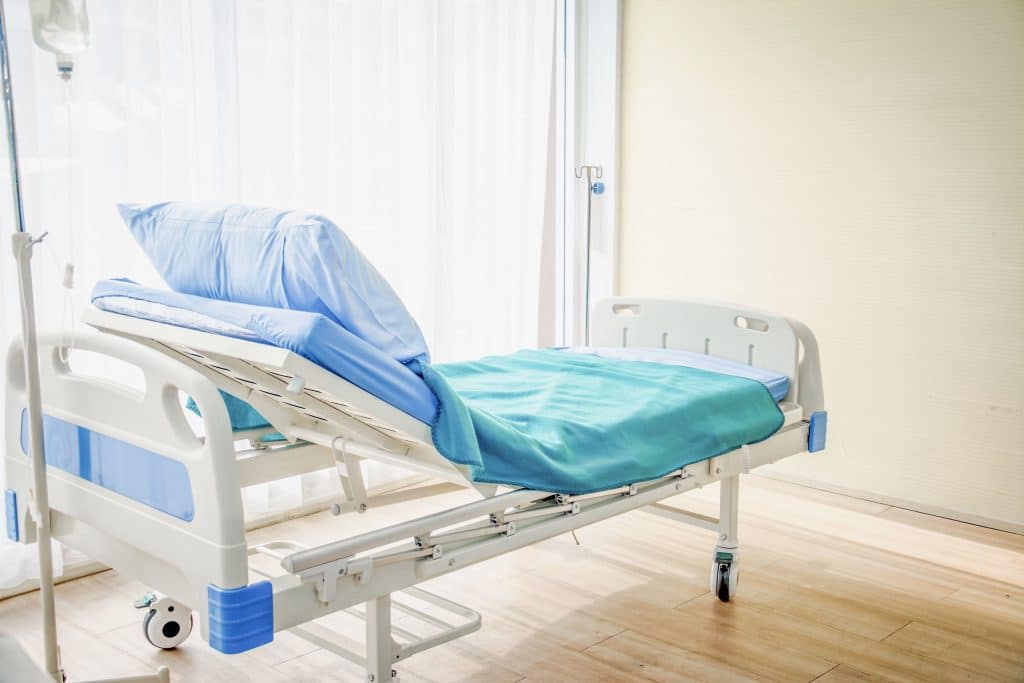 Un lit d'hôpital vide au dossier relevé, face au mur d'une chambre d'hôpital, à côté d'une baie vitrée lumineuse. 