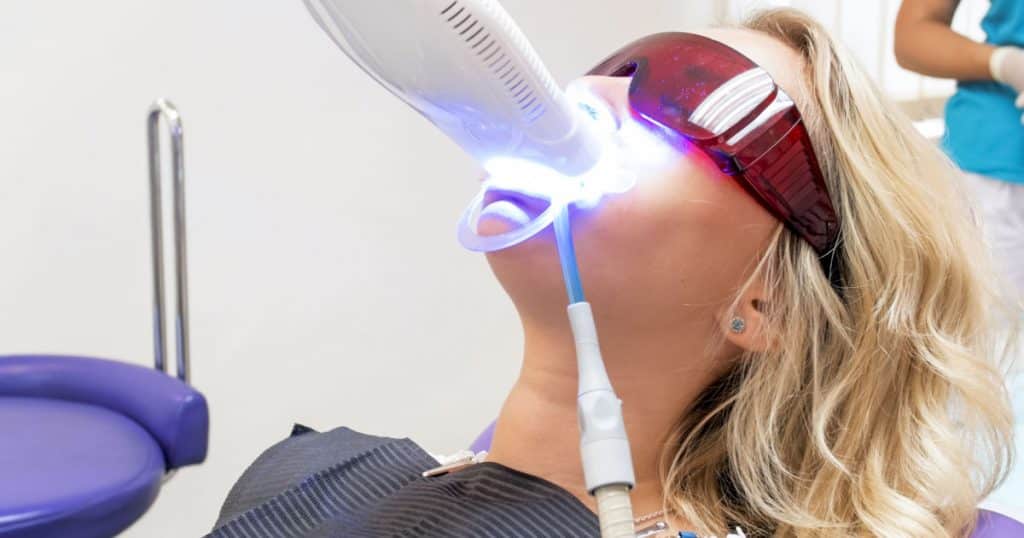 Une femme avec des lunettes de protection se fait blanchir les dents au laser, assise sur un fauteuil de cabinet dentaire.