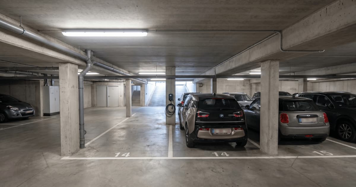 Une borne de recharge montée sur pied est installée sur une place de parking numérotée dans un parking souterrain partagé d'un immeuble résidentiel, avec une voiture électrique branchée et d'autres véhicules garés dans les emplacements voisins.
