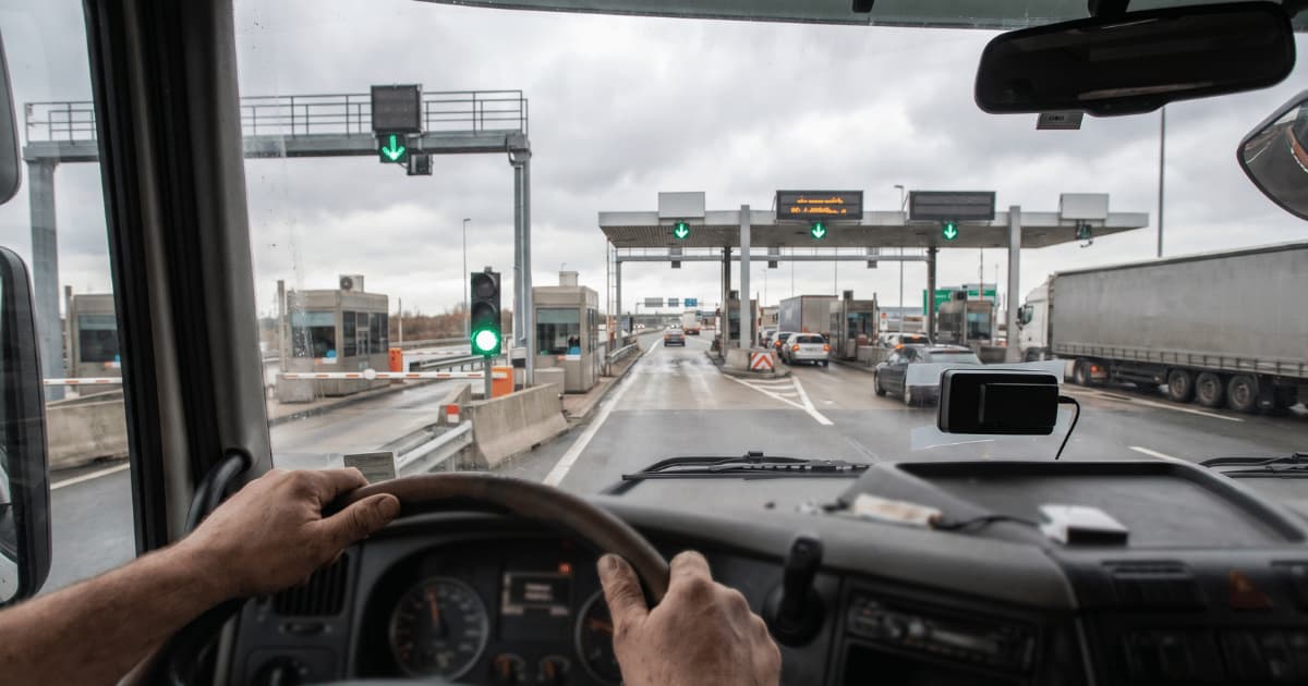 Vue depuis la cabine d'un camion dont le chauffeur tient le volant des deux mains en approchant une barrière de péage autoroutière sous un ciel couvert avec des flèches vertes indiquant le passage libre.