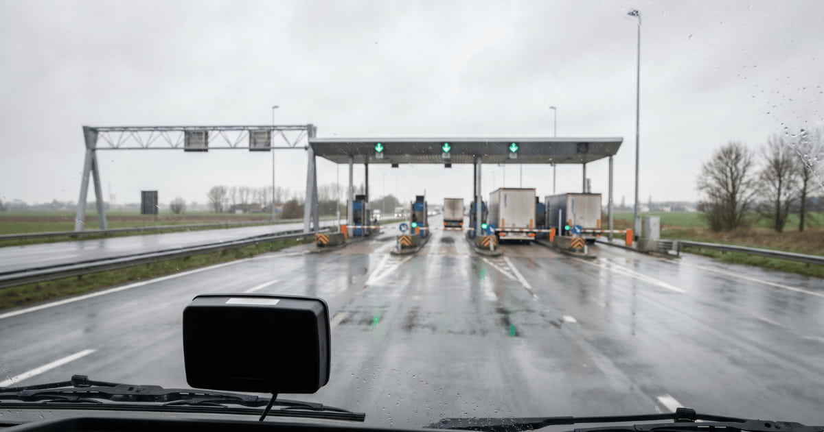 Vue depuis la cabine d'un camion approchant un péage autoroutier par temps de pluie avec des gouttes sur le pare-brise, des feux verts au-dessus des voies et plusieurs poids lourds passant les barrières sur une chaussée mouillée bordée de paysage plat.