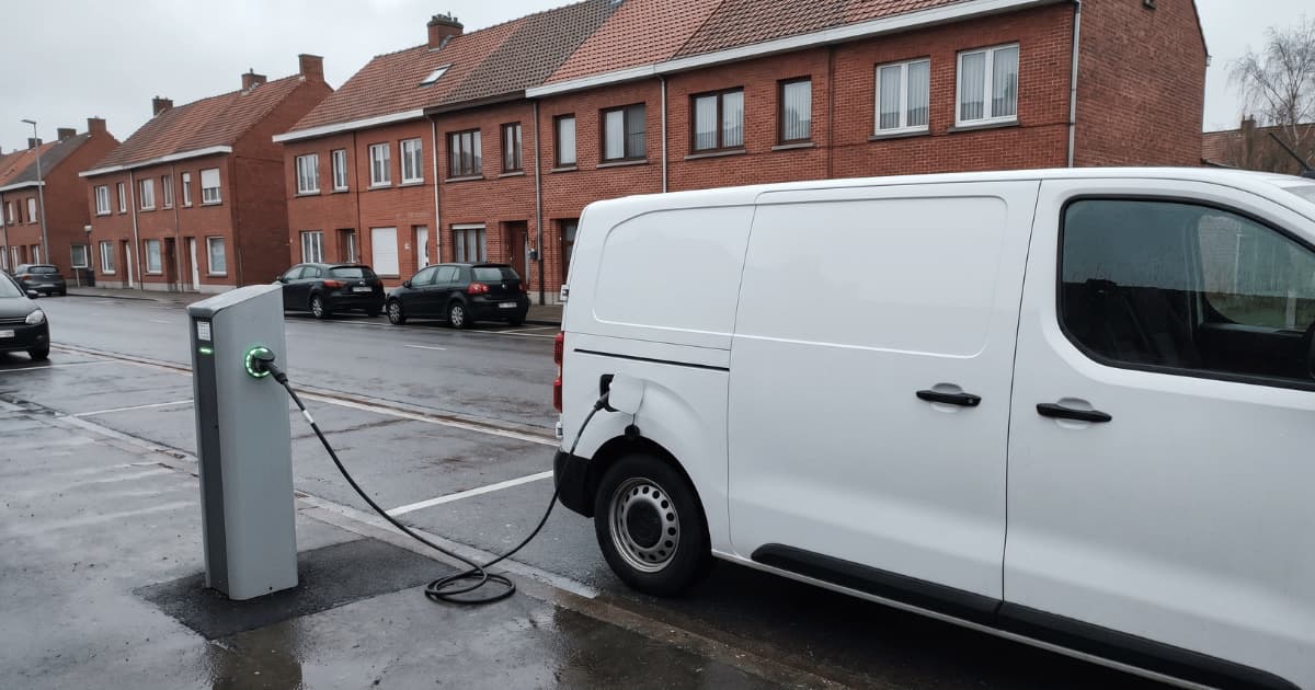 Une camionnette électrique blanche branchée à une borne de recharge publique avec un voyant vert sur un parking de quartier résidentiel belge bordé de maisons mitoyennes en brique rouge sous un ciel gris pluvieux.