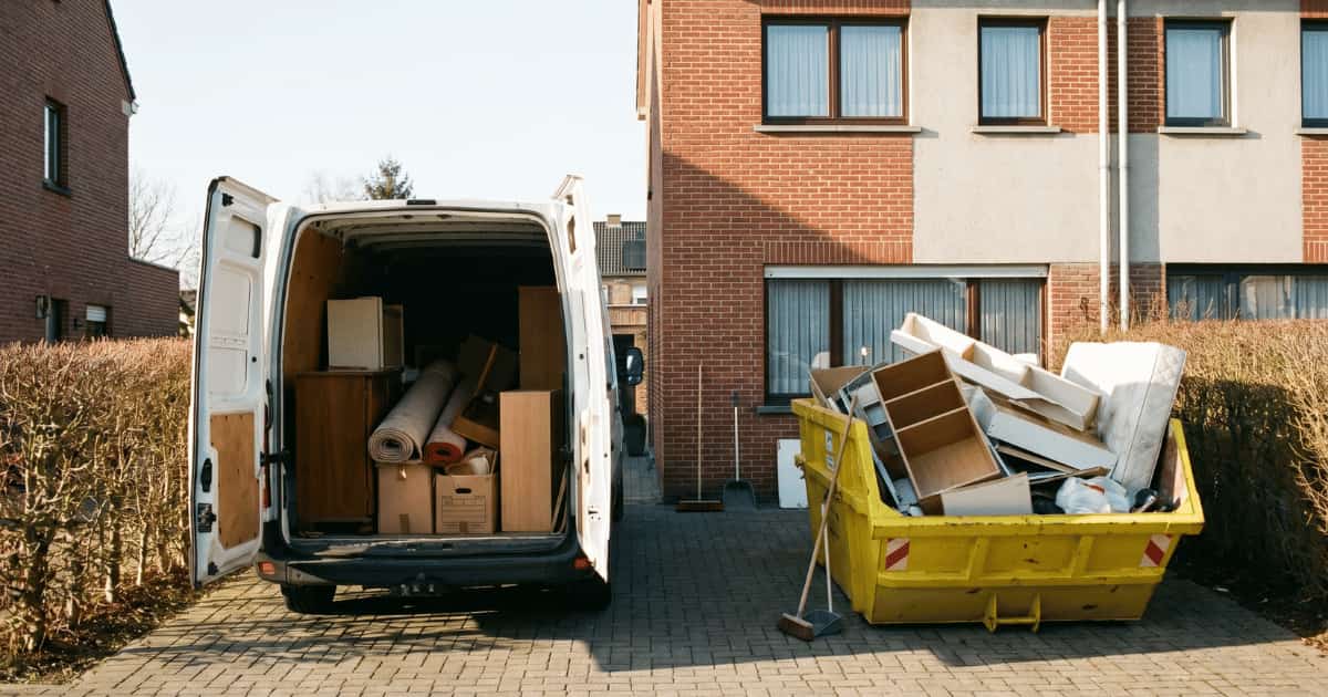 Une camionnette blanche aux portes arrière ouvertes chargée de meubles et un conteneur jaune débordant de mobilier usagé occupent l'allée pavée devant une maison mitoyenne en briques sous un ciel dégagé.
